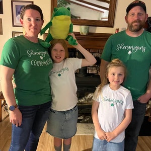May include: A family of four wearing green and white shirts with the text "Shenanigans" and a shamrock. The shirts are a fun way to celebrate St. Patrick's Day.