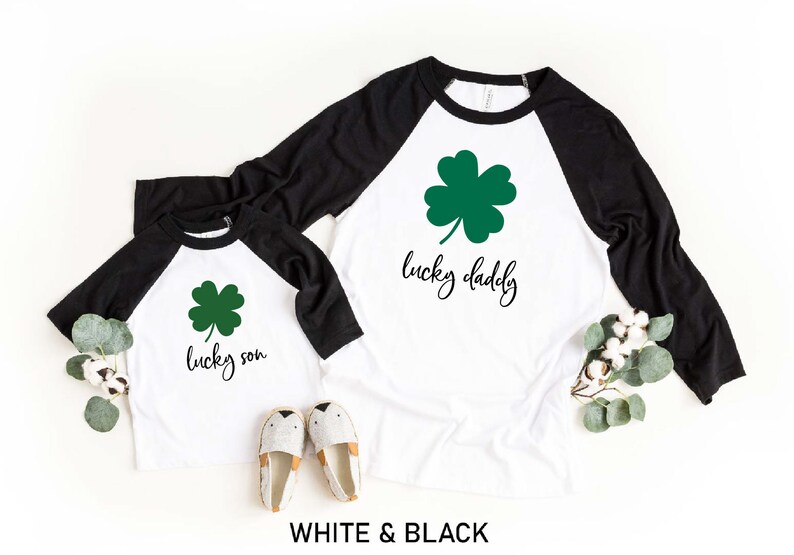 May include: Two white and black baseball shirts with green shamrock designs. The larger shirt says "lucky daddy" and the smaller shirt says "lucky son".