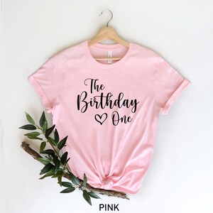 May include: Pink t-shirt with black text that reads "The Birthday One" with a heart symbol.