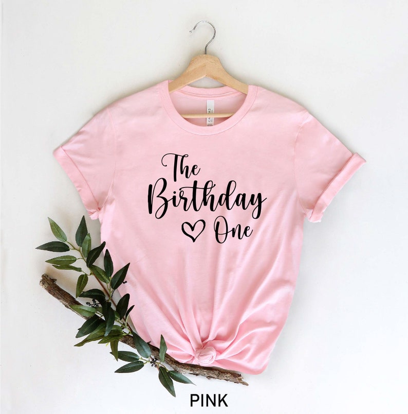 May include: Pink t-shirt with black text that reads "The Birthday One" with a heart symbol.