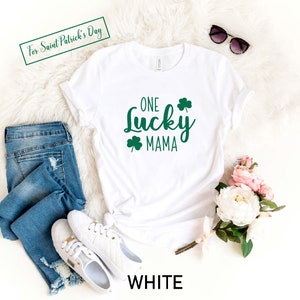 May include: White t-shirt with the text "ONE LUCKY MAMA" in green with shamrock designs. The shirt is laying on a white fuzzy surface with a pair of jeans, white sneakers, sunglasses, and a bouquet of pink and white flowers.