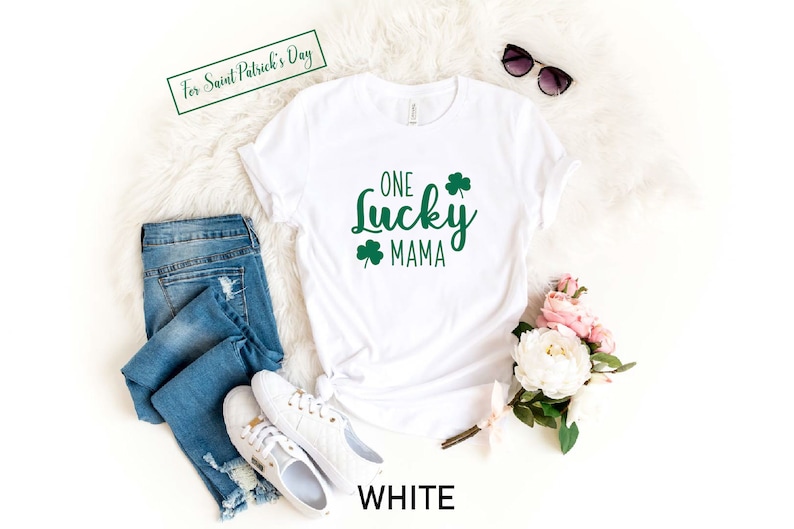 May include: White t-shirt with the text "ONE LUCKY MAMA" in green with shamrock designs. The shirt is laying on a white fuzzy surface with a pair of jeans, white sneakers, sunglasses, and a bouquet of pink and white flowers.