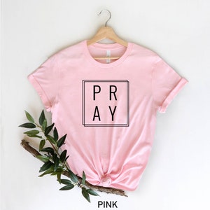 May include: A pink t-shirt with the word "PRAY" in a black square on the front. The shirt is on a hanger and has a rolled up sleeve.