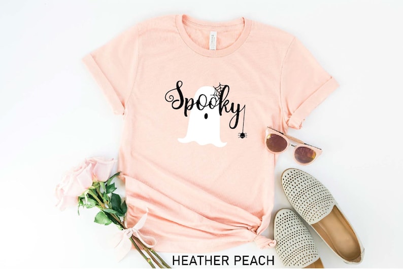 May include: A heather peach colored t-shirt with the text "Spooky" in black cursive font. A white ghost with a black spider web and a spider hanging from it is above the text.