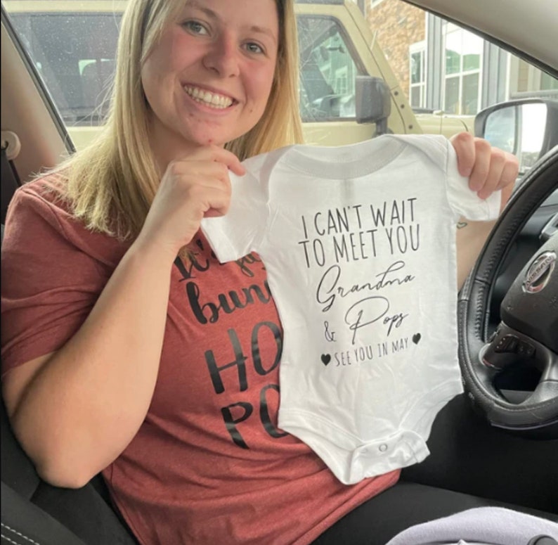 May include: A white onesie with black text that reads "I can't wait to meet you Grandma & Pops See you in May" with a heart on each side of the text.