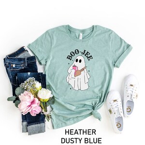 May include: A light blue t-shirt with a ghost holding a pink drink and the text "BOO-JEE" printed on it. The shirt is laid flat on a white background with jeans, flowers, and white sneakers.