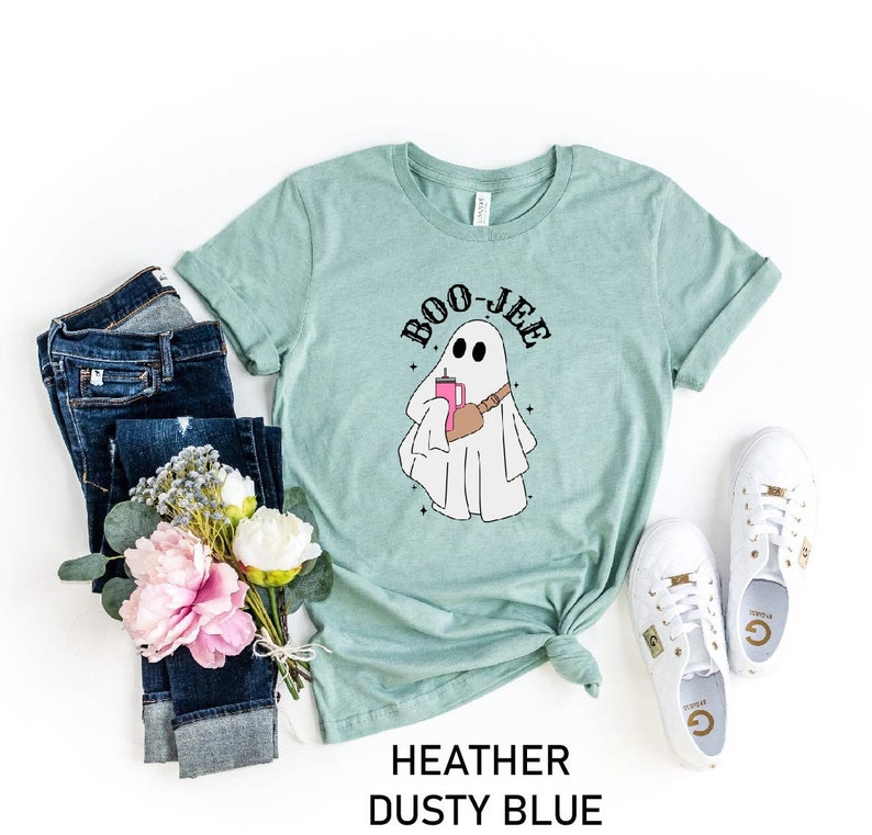 May include: A light blue t-shirt with a ghost holding a pink drink and the text "BOO-JEE" printed on it. The shirt is laid flat on a white background with jeans, flowers, and white sneakers.
