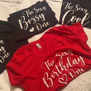 May include: Three red, black, and white t-shirts with a white script design. The red shirt says "The Sexy Birthday One" with a heart. The black shirt says "The Sexy Bossy One" with a heart. The other black shirt says "The Sexy Cool One".