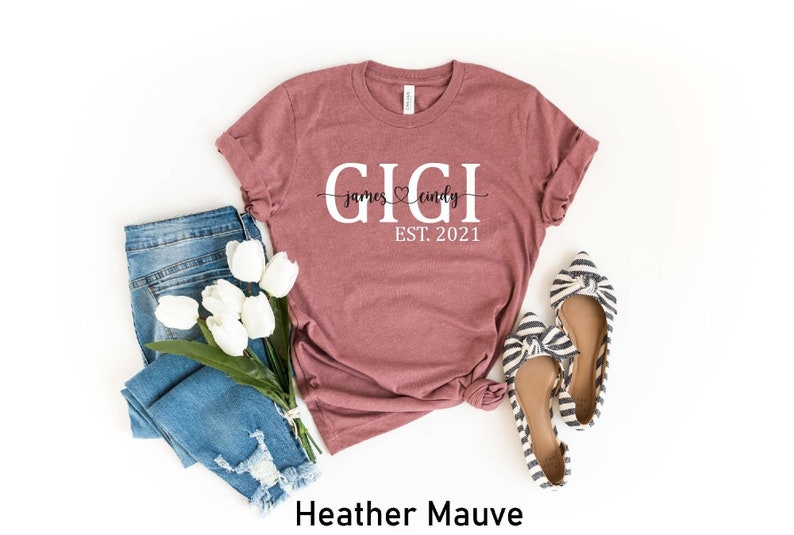 May include: Heather mauve t-shirt with the text "GIGI james &hearts; cindy EST. 2021" printed on it. The shirt is laid flat on a white background with a pair of blue jeans and white flowers.