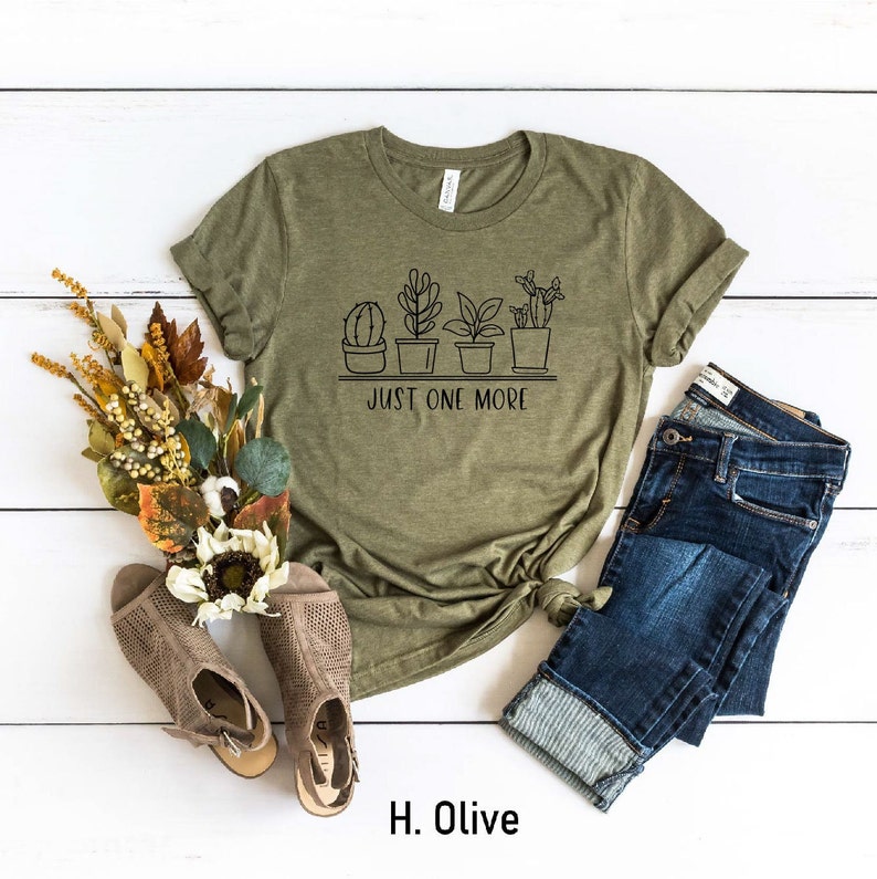 May include: Olive green t-shirt with a graphic of potted plants and the text "Just One More".