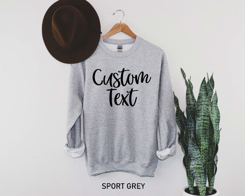 May include: A gray sweatshirt with the text "Custom Text" in black lettering. The sweatshirt is hanging on a wooden hanger against a white wall.