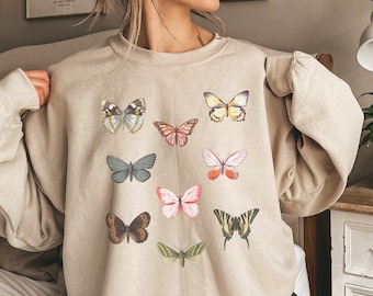 May include: A beige sweatshirt with a crew neck and puffed sleeves features a pattern of colorful butterflies. The butterflies are in various sizes and positions, creating a whimsical and playful design.