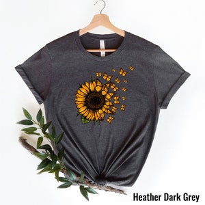 K&ouml;nnte beinhalten: Ein dunkelgraues T-Shirt mit einem Sonnenblumen-Motiv. Die Sonnenblume hat gelbe Bl&uuml;tenbl&auml;tter und eine braune Mitte. Schmetterlinge fliegen aus der Sonnenblume.