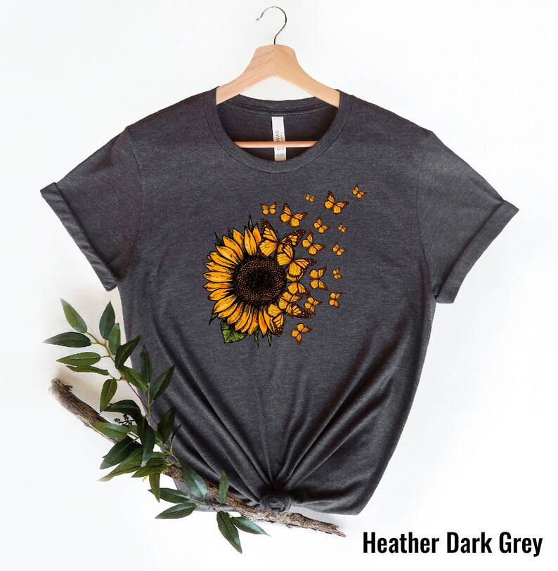 K&ouml;nnte beinhalten: Ein dunkelgraues T-Shirt mit einem Sonnenblumen-Motiv. Die Sonnenblume hat gelbe Bl&uuml;tenbl&auml;tter und eine braune Mitte. Schmetterlinge fliegen aus der Sonnenblume.