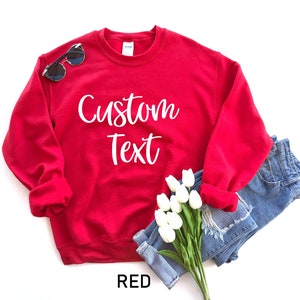 May include: Red sweatshirt with the text "Custom Text" in white lettering. The sweatshirt is folded and displayed with a pair of sunglasses and a bouquet of white flowers.