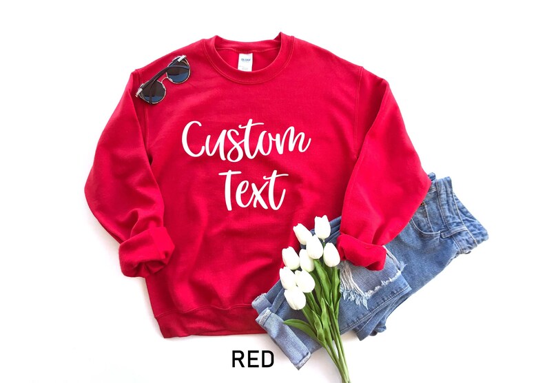 May include: Red sweatshirt with the text "Custom Text" in white lettering. The sweatshirt is folded and displayed with a pair of sunglasses and a bouquet of white flowers.