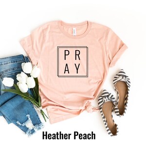 May include: A heather peach colored t-shirt with the word "PRAY" in a black square frame. The shirt is laid flat on a white background with a pair of blue jeans, white tulips, and striped flats.