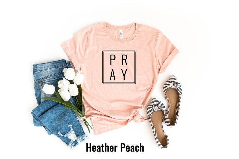 May include: A heather peach colored t-shirt with the word "PRAY" in a black square frame. The shirt is laid flat on a white background with a pair of blue jeans, white tulips, and striped flats.
