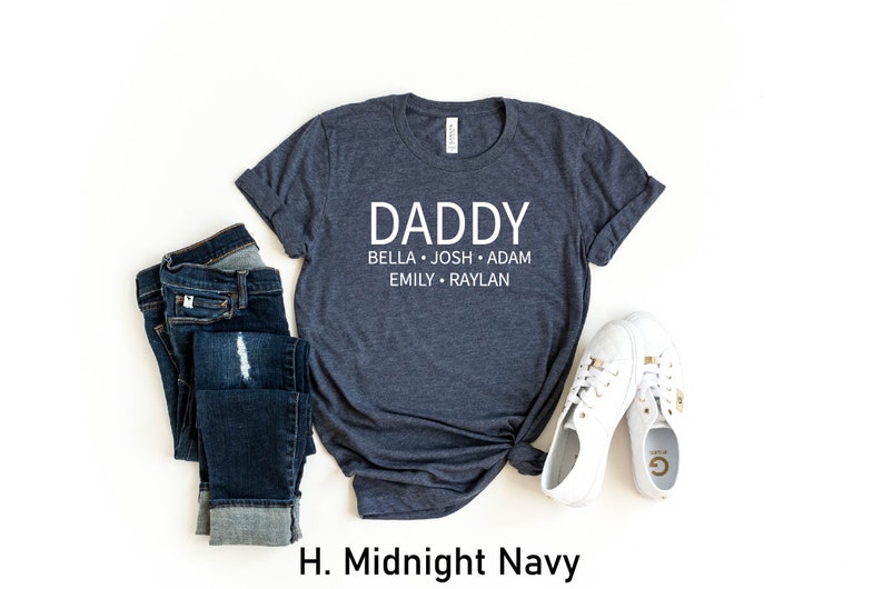May include: A dark blue t-shirt with the text "DADDY" and the names "BELLA, JOSH, ADAM, EMILY, RAYLAN" printed in white. The shirt is displayed on a white background with a pair of blue jeans and white sneakers.