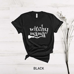 Może przedstawiać: Czarny T-shirt z napisem "witchy mama" w białym kolorze i białą sylwetką miotły.
