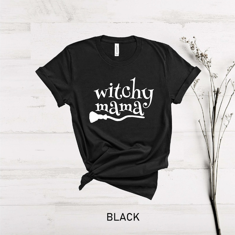Może przedstawiać: Czarny T-shirt z napisem "witchy mama" w białym kolorze i białą sylwetką miotły.