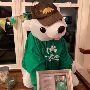 May include: A white polar bear wearing a green hoodie with a shamrock and the word "Michigan" on it. The bear is also wearing a brown baseball cap with a gold bear and marijuana leaf design. The bear is sitting on a wooden table with a framed picture of a person wearing a similar green hoodie. The picture has a poem titled "A Celebration of Life" on it.