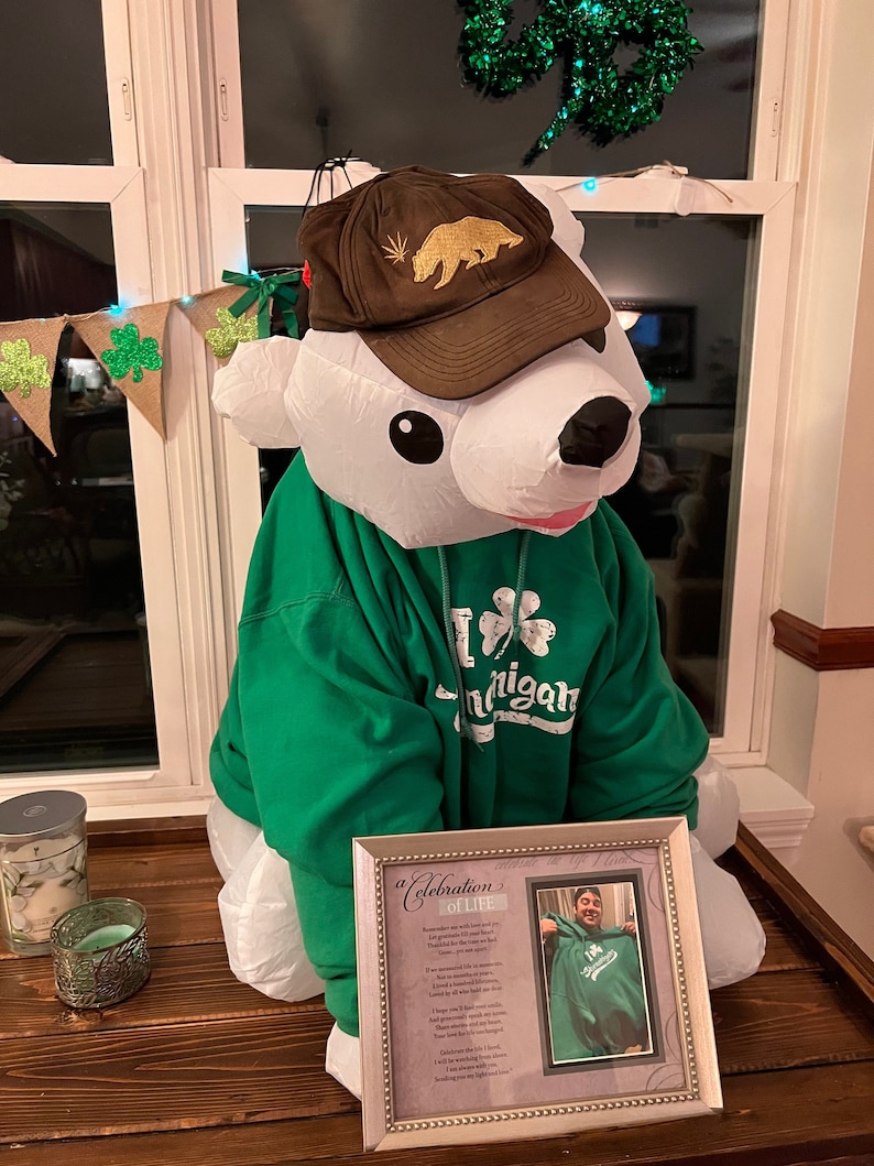 May include: A white polar bear wearing a green hoodie with a shamrock and the word "Michigan" on it. The bear is also wearing a brown baseball cap with a gold bear and marijuana leaf design. The bear is sitting on a wooden table with a framed picture of a person wearing a similar green hoodie. The picture has a poem titled "A Celebration of Life" on it.