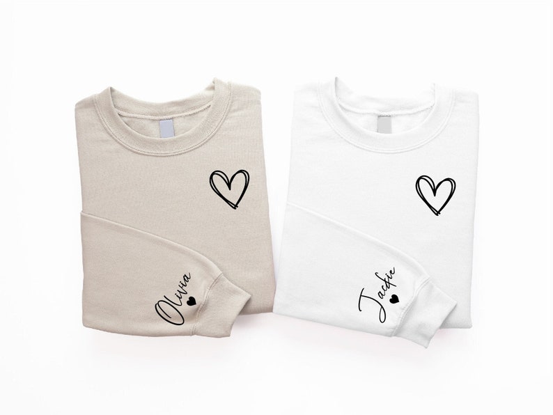 Peut inclure: Deux sweats beige et blanc avec un c&oelig;ur et un nom sur chacun. Le sweat beige a "Olivia" sur la manche et le sweat blanc a "Jackie" sur la manche.