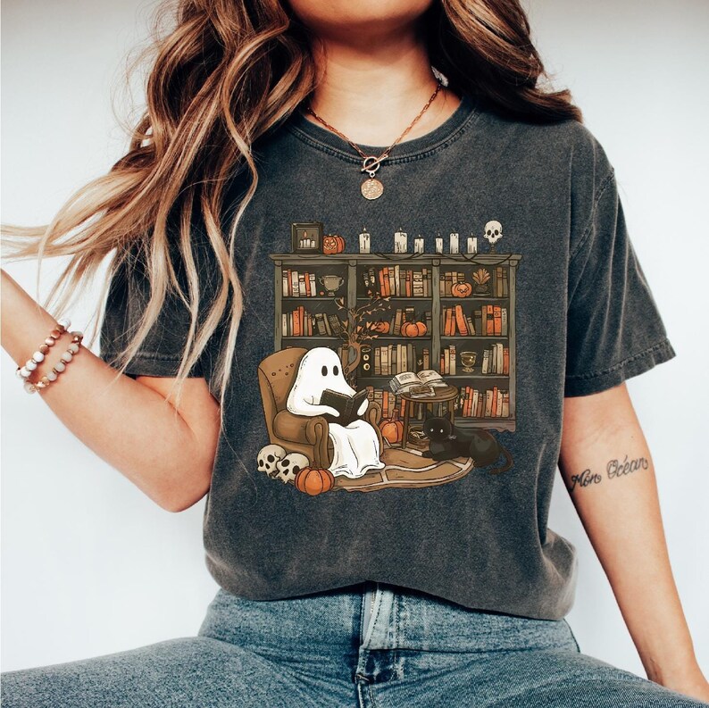 May include: A dark gray t-shirt with a graphic of a ghost reading a book in a chair in front of a bookshelf filled with books and pumpkins. The ghost is white and the bookshelf is brown. The text "More Goblins" is on the right sleeve of the shirt.