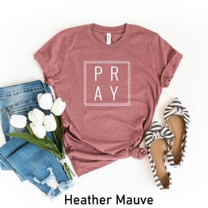 May include: A heather mauve t-shirt with the word "PRAY" in a white box on the front. The shirt is laid out on a white background with a pair of blue jeans, a bouquet of white tulips, and a pair of striped flats.