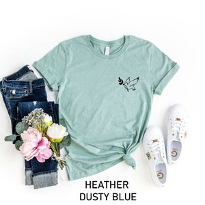May include: A heather dusty blue t-shirt with a black dove graphic holding an olive branch. The shirt is styled with blue jeans, white sneakers, and pink flowers.