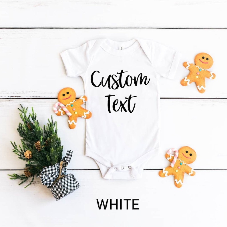 May include: White baby bodysuit with the text "Custom Text" printed in black. The bodysuit is surrounded by gingerbread cookies on a white wooden background.