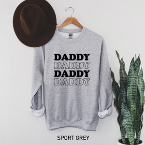 K&ouml;nnte beinhalten: Ein grauer Pullover mit dem Wort "Daddy", das viermal in Schwarz und Wei&szlig; geschrieben ist. Der Text ist in einer Retro-Schriftart. Der Pullover h&auml;ngt an einem Kleiderb&uuml;gel und hat einen wei&szlig;en Hintergrund.