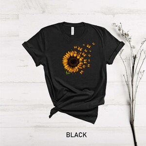 K&ouml;nnte beinhalten: Schwarzes T-Shirt mit einem Sonnenblumen-Motiv und Schmetterlingen, die davon fliegen. Die Sonnenblume ist gelb und braun, und die Schmetterlinge sind orange.