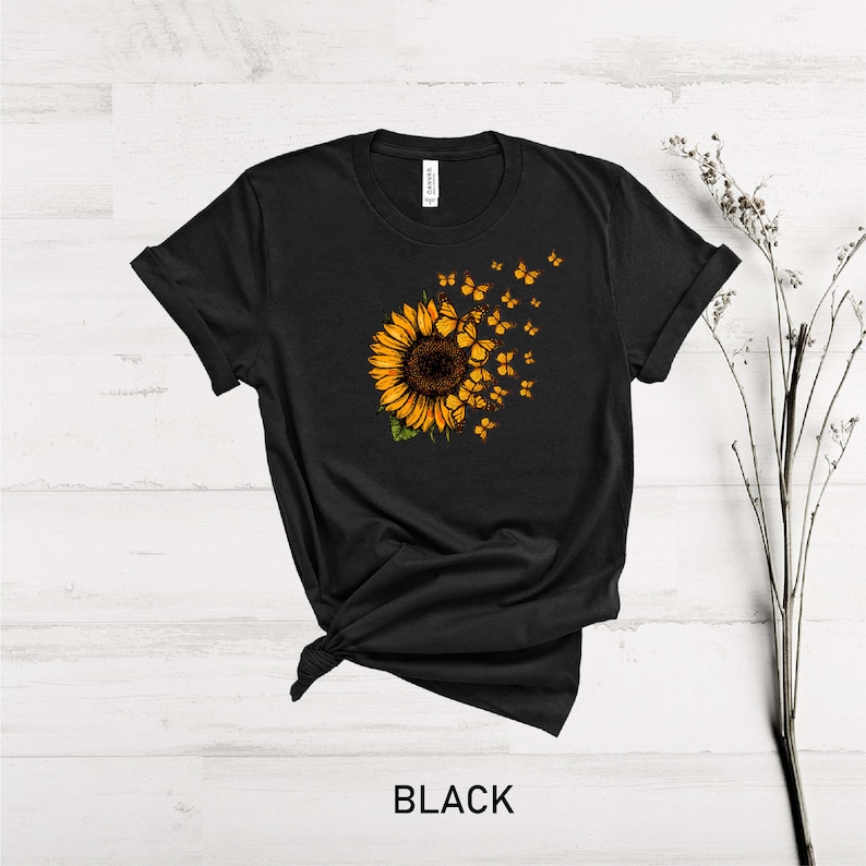 K&ouml;nnte beinhalten: Schwarzes T-Shirt mit einem Sonnenblumen-Motiv und Schmetterlingen, die davon fliegen. Die Sonnenblume ist gelb und braun, und die Schmetterlinge sind orange.
