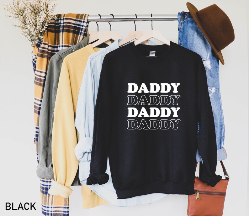 K&ouml;nnte beinhalten: Schwarzer Pullover mit dem Text "DADDY", der viermal in einer gestapelten Retro-Schriftart wiederholt wird.