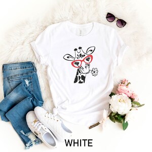 May include: White t-shirt featuring a black and white illustration of a giraffe wearing red glasses and holding a flower. The shirt is laid flat on a white fluffy surface with a pair of sunglasses, a pair of jeans, and a bouquet of flowers.