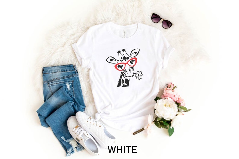 May include: White t-shirt featuring a black and white illustration of a giraffe wearing red glasses and holding a flower. The shirt is laid flat on a white fluffy surface with a pair of sunglasses, a pair of jeans, and a bouquet of flowers.