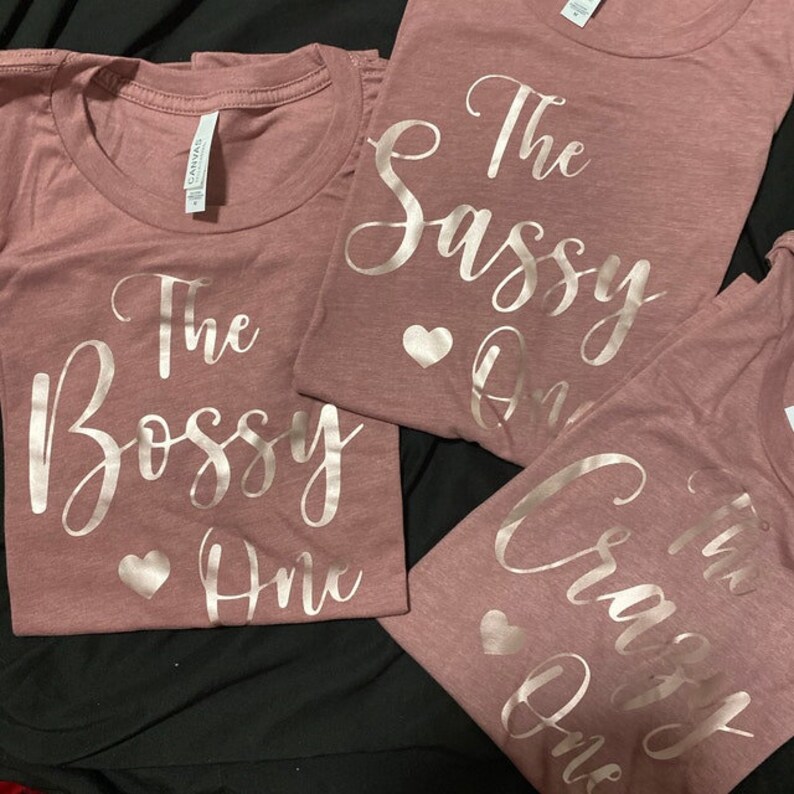 May include: Three pink t-shirts with rose gold foil lettering. The shirts say "The Bossy One", "The Sassy One", and "The Crazy One".