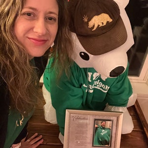 May include: A woman in a green shirt poses with a large inflatable polar bear wearing a brown baseball cap with a gold bear and marijuana leaf logo. The bear is wearing a green sweatshirt with the word "Michigan" on it. A framed picture with a poem titled "A Celebration of Life" is in the foreground.