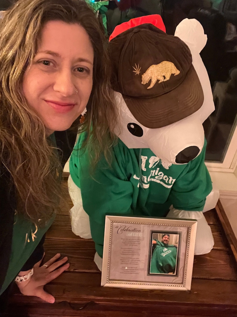 May include: A woman in a green shirt poses with a large inflatable polar bear wearing a brown baseball cap with a gold bear and marijuana leaf logo. The bear is wearing a green sweatshirt with the word "Michigan" on it. A framed picture with a poem titled "A Celebration of Life" is in the foreground.