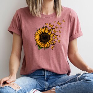 K&ouml;nnte beinhalten: Ein pinkfarbenes T-Shirt mit einem Sonnenblumen-Motiv und Schmetterlingen, die davonfliegen. Die Sonnenblume ist gelb mit einer braunen Mitte und gr&uuml;nen Bl&auml;ttern. Die Schmetterlinge sind gelb mit schwarzen Markierungen.