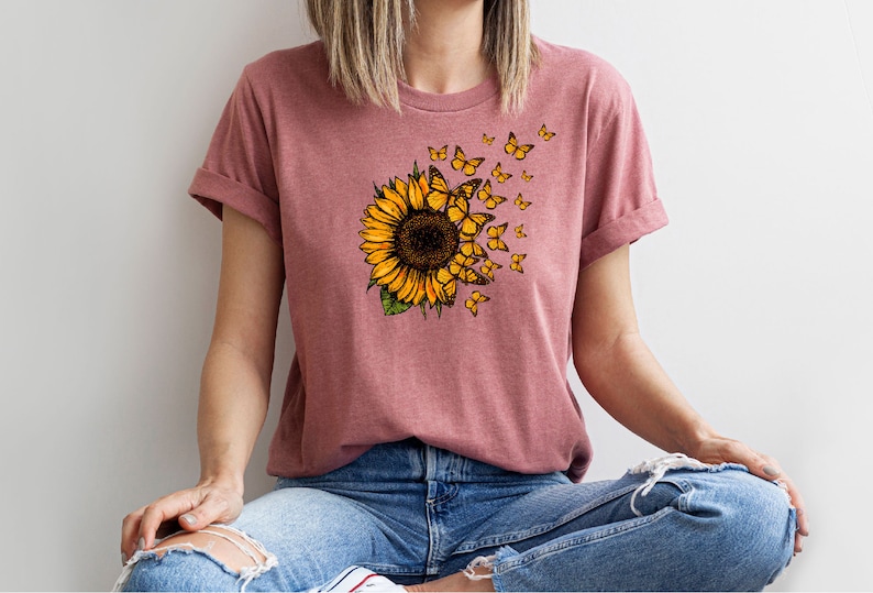K&ouml;nnte beinhalten: Ein pinkfarbenes T-Shirt mit einem Sonnenblumen-Motiv und Schmetterlingen, die davonfliegen. Die Sonnenblume ist gelb mit einer braunen Mitte und gr&uuml;nen Bl&auml;ttern. Die Schmetterlinge sind gelb mit schwarzen Markierungen.