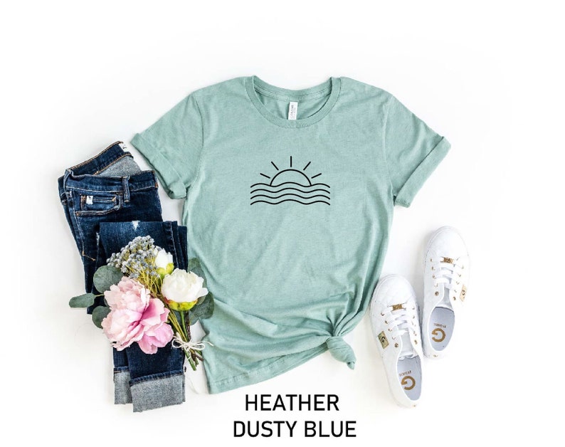 May include: A heather dusty blue t-shirt featuring a simple black graphic of a sun setting over wavy lines. The shirt is styled with blue jeans, a bouquet of pink flowers, and white trainers.
