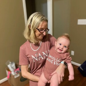 May include: A woman wearing a pink shirt with the text "Nana" on it is holding a baby wearing a pink onesie with the text "Nana's lil sweet pea".