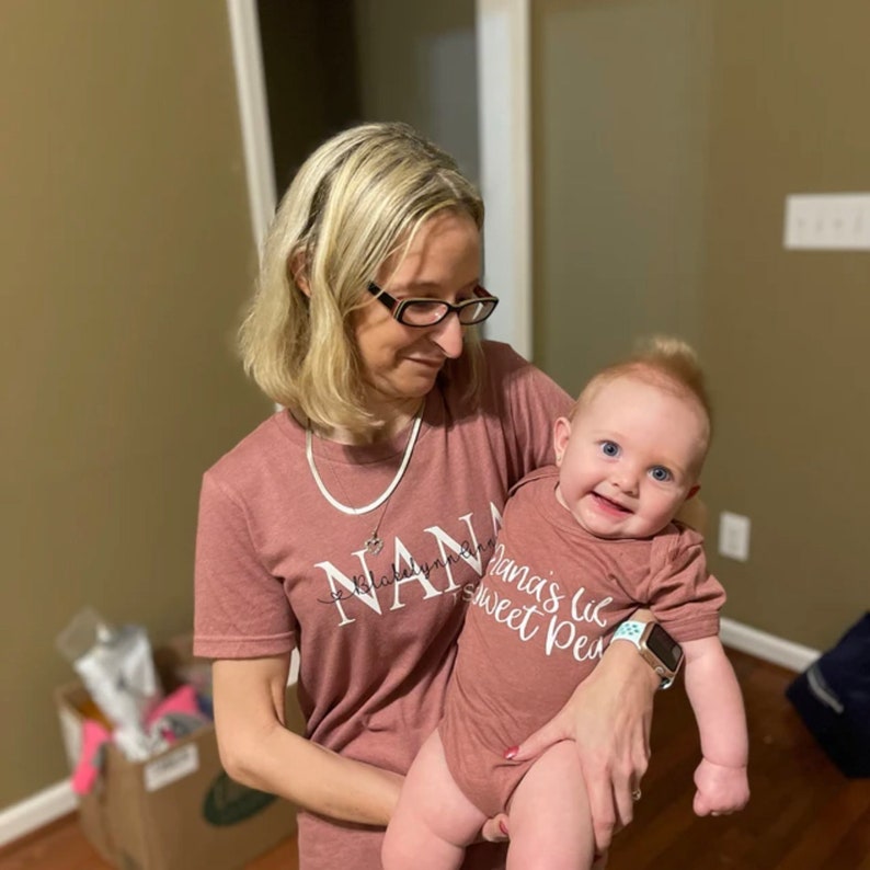 May include: A woman wearing a pink shirt with the text "Nana" on it is holding a baby wearing a pink onesie with the text "Nana's lil sweet pea".