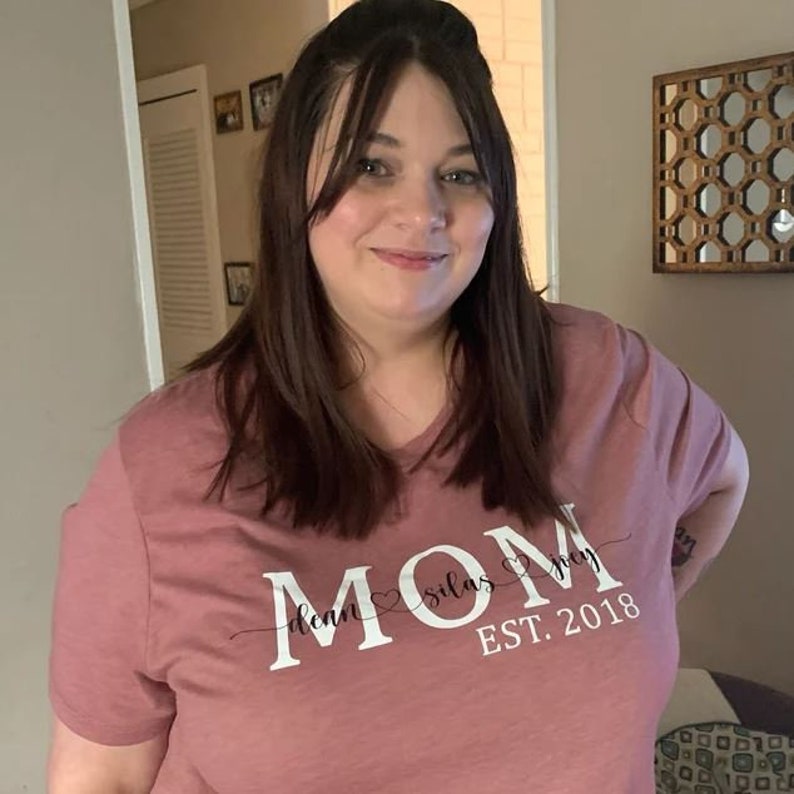 May include: A woman wearing a mauve t-shirt with the text "MOM" in white with the words "dean", "silas", and "grey" written in cursive script with hearts between each name. The text "EST. 2018" is written below the names.
