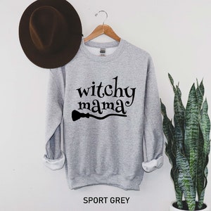 May include: A gray sweatshirt with the text "witchy mama" and a black silhouette of a broom underneath. The sweatshirt is on a hanger against a white wall.