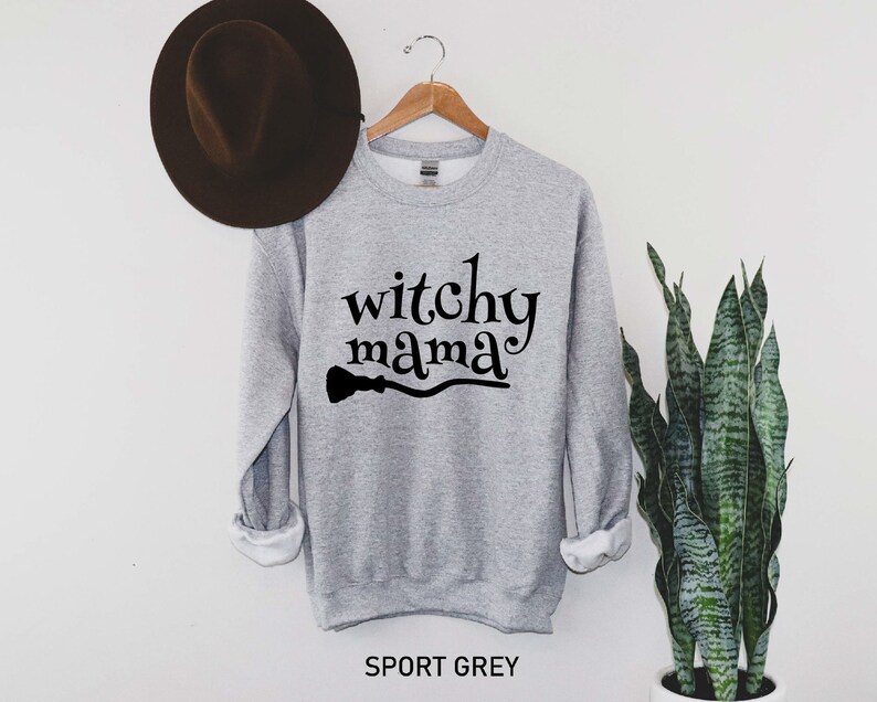 May include: A gray sweatshirt with the text "witchy mama" and a black silhouette of a broom underneath. The sweatshirt is on a hanger against a white wall.