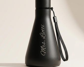 May include: A matte black water bottle with a wide base and a narrow neck, featuring a screw-top lid and a black carrying strap. The text Mrs. Liotti is engraved in a white cursive script on the front of the bottle, which has a smooth finish.